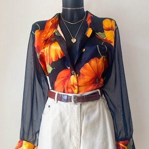 Op de afbeelding: Een overhemd met lange mouwen en knoopsluiting, voorzien van een zwarte basis met grote oranje en gele bloemmotieven, transparante zwarte mouwen en bijpassende manchetten. Het overhemd zit in een gebroken witte broek met een bruine leren riem.