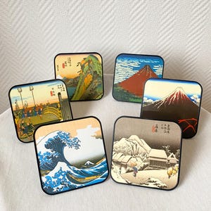 Peut inclure: Ensemble de six dessous de verre carrés avec des bordures noires, chacun orné d'une estampe japonaise différente. Les images représentent des paysages variés, notamment des montagnes et des vagues. Ces dessous de verre sont pour les boissons.