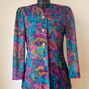 Peut inclure: Blazer vintage avec un motif floral vibrant en teal, violet, rose et vert. La veste à manches longues est ornée de boutons dorés et d'un col classique. Le blazer est présenté sur un mannequin.