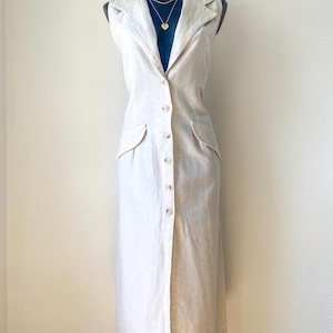 Peut inclure: Une robe midi sans manches de couleur blanc cassé avec un col à revers cranté, une fermeture boutonnée sur toute la longueur à l'avant et deux poches latérales à rabat. Le vêtement est présenté sur un mannequin sombre, montrant une coupe structurée.