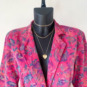 May include: A bright pink corduroy blazer featuring a multi-colored floral pattern in shades of purple, blue, and orange. The blazer is styled with three layered gold-tone necklaces, including a choker, a chain, and a heart-shaped pendant.
