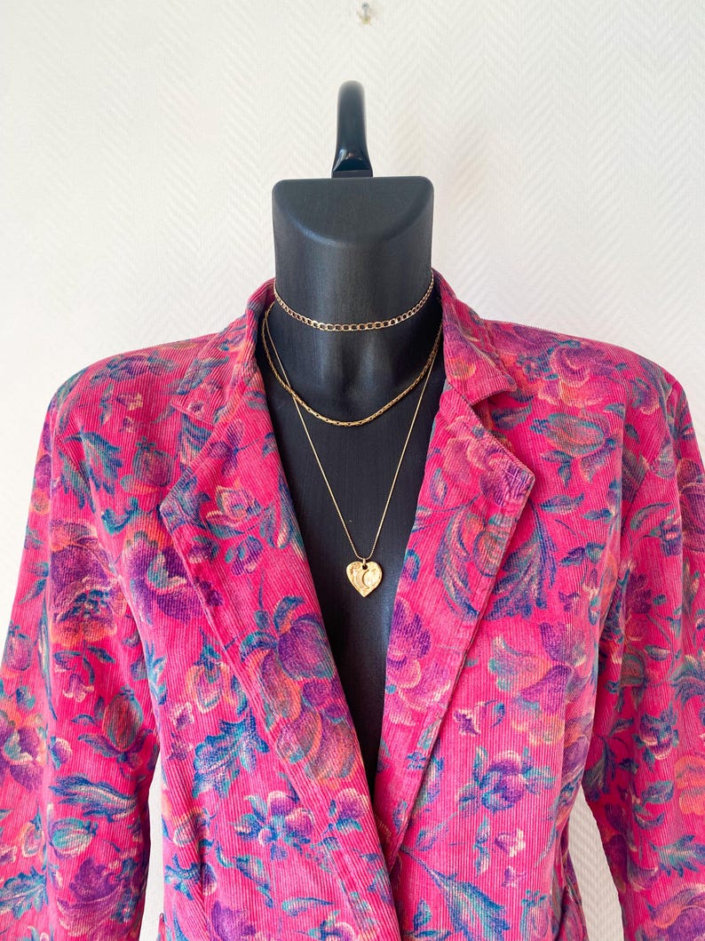 May include: A bright pink corduroy blazer featuring a multi-colored floral pattern in shades of purple, blue, and orange. The blazer is styled with three layered gold-tone necklaces, including a choker, a chain, and a heart-shaped pendant.