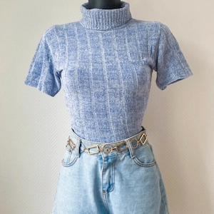 Peut inclure: Un haut bleu clair à col roulé et manches courtes côtelé, associé à un short en jean délavé. Une ceinture chaîne dorée à maillons géométriques accessoirise le short. L'ensemble est présenté sur un mannequin.