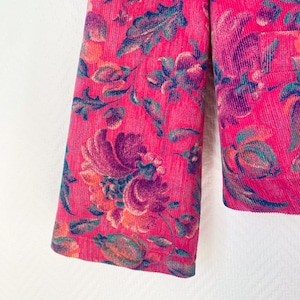 May include: A bright pink corduroy jacket featuring a prominent floral pattern in shades of deep purple, teal, and orange. The garment has a textured ribbed surface and a visible pocket on the right side, showcasing a dense botanical print.