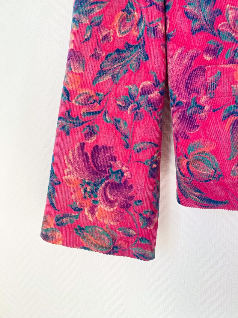 May include: A bright pink corduroy jacket featuring a prominent floral pattern in shades of deep purple, teal, and orange. The garment has a textured ribbed surface and a visible pocket on the right side, showcasing a dense botanical print.