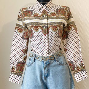 Peut inclure: Chemise blanche à motifs cachemire rouges, verts et dorés. Elle est associée à un short en jean bleu clair et une ceinture chaîne dorée. L'ensemble est présenté sur un mannequin noir.