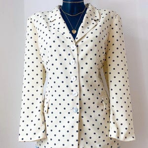 Peut inclure: Blazer crème à pois noirs. Le blazer a un col cranté classique, une fermeture boutonnée et deux poches avant. Le vêtement est présenté sur un mannequin noir, associé à un short en jean et des colliers dorés.