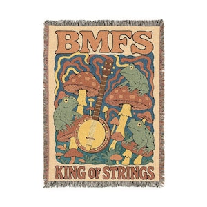 May include: Woven throw blanket with a whimsical design. The tan blanket features the text "BMFS" and "KING OF STRINGS" in brown. The design includes a banjo, mushrooms, and green frogs against a blue and yellow background.