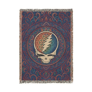 May include: A woven throw blanket featuring a central skull and lightning bolt design within a circular pattern. The blanket has a blue background with red and green accents, and a fringed edge.