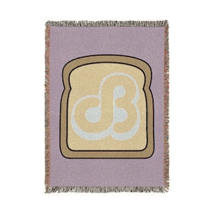 May include: A woven throw blanket featuring a light purple background and a central graphic of a slice of toast. The toast has a dark brown crust, a tan center, and a stylized, light blue, interlocking letter design printed on the bread surface.