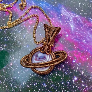 May include: A wire-wrapped pendant shaped like a planet with rings, featuring a round, banded purple and white stone at its center. The pendant is crafted from copper-colored wire and hangs from a matching copper-colored metal chain link necklace.