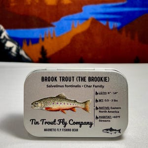 May include: A rectangular silver metal tin featuring a graphic of a Brook Trout with the text Brook Trout (The Brookie), Salvelinus fontinalis, Char Family, Tin Trout Fly Company, and Magnetic Fly Fishing Gear, plus data on length, weight, and habitat.