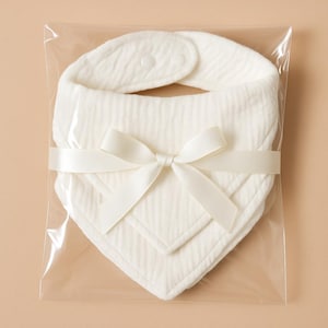 May include: A set of two off-white baby bibs, made of soft, textured fabric, are neatly packaged in a clear plastic bag. The bibs are secured with a matching ivory ribbon tied in a bow. The bibs have a snap closure.