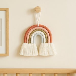 May include: A handmade rainbow wall hanging with a peach, pink, gold, and gray color scheme. The rainbow is made of yarn and has a white fringe at the bottom. It is hanging on a wall with a wooden peg.