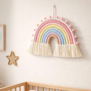 May include: A colorful, handmade rainbow wall hanging. The rainbow features pastel shades of pink, yellow, blue, and tan, with a fringe at the bottom and pink pom-poms along the top. It is a decorative piece for a nursery or child's room.