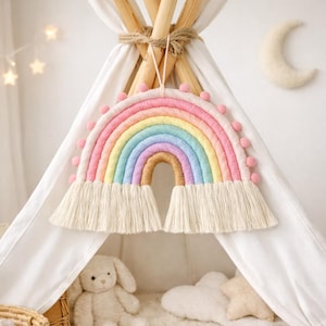 May include: A colorful rainbow wall hanging with pastel shades of pink, yellow, blue, and tan. The rainbow is adorned with pink pom-poms and fringed edges. It is suspended in front of a white teepee, creating a whimsical nursery decor.