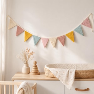 May include: A fabric bunting banner featuring triangular flags in muted shades of cream, dusty pink, mustard yellow, and sage green, suspended on a white cord. The flags are made of a textured, crinkled fabric and hang in a decorative scalloped arc.