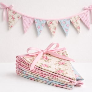 May include: A stack of floral patterned fabric pennants with pink ribbon tied on top. The pennants are in shades of pink, blue, and cream. A matching banner of pennants hangs in the background, creating a cohesive decorative theme.