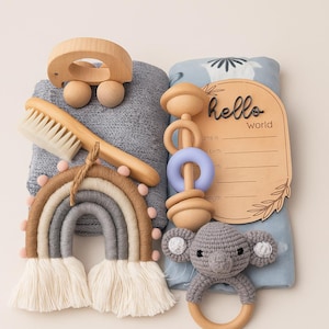 May include: A collection of baby items including a wooden car, a soft brush, a rainbow wall hanging, a wooden rattle with a blue ring, a crocheted koala rattle, and a baby blanket with a "hello world" sign.