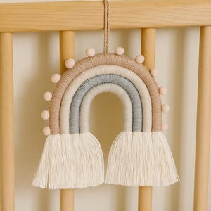 May include: A decorative wall hanging shaped like a rainbow, constructed from wrapped yarn in shades of tan, cream, and light blue. The arch features small, light pink pom-poms along the outer edge and two long, cream-colored yarn tassels.