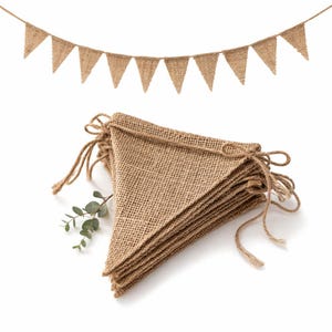 May include: A set of burlap pennant banners. One banner is strung with triangular flags, and another is a stack of folded flags. The flags are a natural tan color, and the string is a matching brown. A sprig of green leaves is next to the stack of flags.