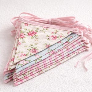 May include: A stack of pink and floral fabric bunting flags with a pink ribbon. The flags feature a variety of patterns, including pink roses on a cream background, light blue fabric, and pink stripes. The bunting is ideal for decorating.