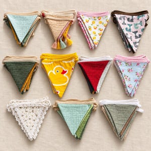 May include: A collection of eleven triangular fabric bunting flags arranged in three rows. The flags feature various patterns including solid colors, floral prints, a yellow duck motif, and a fox print, with one white crocheted lace flag.