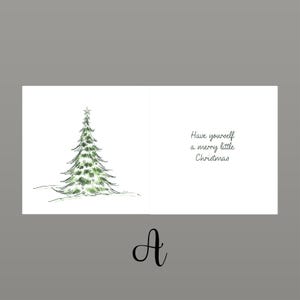 May include: A Christmas card featuring a watercolour illustration of a green Christmas tree on the left. The right side displays the text "Have yourself a merry little Christmas". The card is open and set against a grey backdrop.