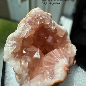 May include: A pale pink crystal geode with translucent, angular formations inside a hollow cavity. The specimen features text stating Size: 22.4x17.5x15.29mm approx and Origin: Argentina. The exterior is a rough, light-colored mineral crust.