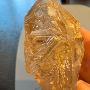 May include: A translucent, amber-colored crystal with a pointed top and rough, textured surfaces. The crystal is held in a hand, showcasing its natural, irregular shape and internal patterns. The gemstone appears to be a raw, uncut specimen.