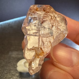 May include: A translucent, light brown crystal with a faceted surface. The crystal has a rough, natural texture and is held in a person's hand. The crystal is a mineral specimen.