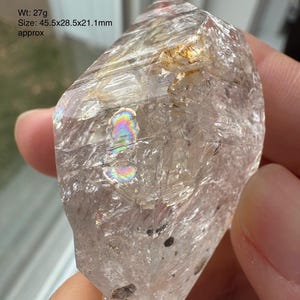 May include: A clear quartz crystal with a weight of 27 grams. The crystal measures approximately 1.8 x 1.1 x 0.8 inches. The surface of the crystal has a textured appearance with iridescent patches.