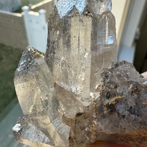 May include: A clear quartz crystal cluster with multiple points. The crystal has a translucent appearance with some areas of grey and black inclusions. The light catches the facets, creating a sparkling effect. The crystal is a natural mineral specimen.