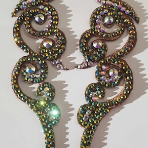 May include: Two decorative embellishments with a symmetrical design. They feature a swirling, openwork pattern in a copper-toned material, densely covered with iridescent, multi-coloured rhinestones. Large, clear rhinestones add sparkle to the design.