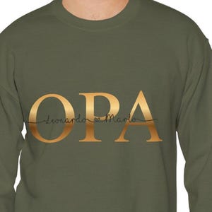 Peut inclure: Un sweat-shirt vert olive avec le mot "OPA" en lettres dorées. En dessous, le texte "Leonardo & Marlo" est écrit en script. Le sweat-shirt est de style col rond.
