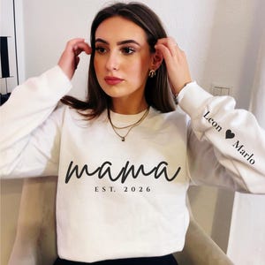 Puede incluir: Sudadera blanca de manga larga con la palabra "mama" en escritura negra y "EST. 2026" debajo. La manga tiene los nombres "Leon" y "Marlo" con un símbolo de corazón. La sudadera la lleva una persona.