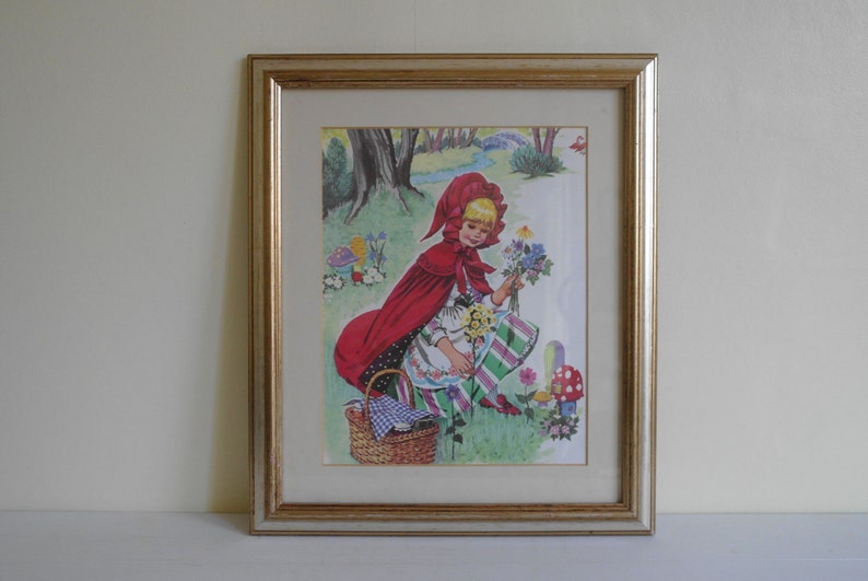 Framed book print of Little Red Riding Hood collecting flowers in the wood, present in a gold frame with a cream mount. Measures approx 35cm by 30cm by 2cm