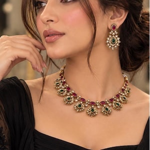 May include: A gold-toned jewellery set featuring a necklace, drop earrings, and a maang tikka. Each piece is adorned with oval-shaped ruby red and teardrop-shaped emerald green stones, surrounded by small white crystals and intricate metalwork.