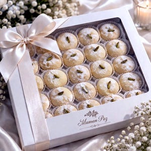 May include: A square white gift box containing twenty-four round, powdered sugar-coated cookies, each topped with a single green pistachio. The box features a large, pale pink satin ribbon bow and the text Maman Paz printed on the front corner.