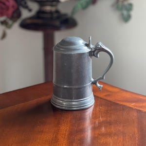 May include: A pewter beer stein with a hinged lid and ornate handle. The cylindrical body of the stein has a textured surface. The handle is curved and detailed. The stein is sitting on a wooden table.
