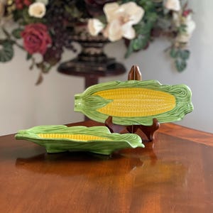 May include: Two ceramic corn-shaped serving dishes featuring a bright yellow cob center surrounded by textured green husks. One dish is positioned horizontally on the surface, while the other is displayed upright on a small wooden stand.