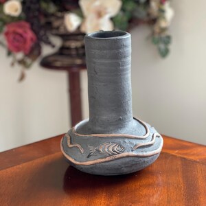 May include: A handcrafted, dark gray ceramic vase with a bulbous base and a cylindrical neck. The base features a raised, light brown design with a fish motif. The vase is displayed on a wooden surface, showcasing its unique, artistic design.
