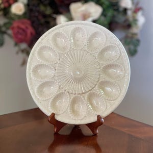 May include: A speckled, cream-coloured ceramic devilled egg serving platter. The round platter has 12 egg-shaped indentations around the edge and a textured, sunburst design in the centre. It is displayed on a wooden stand.