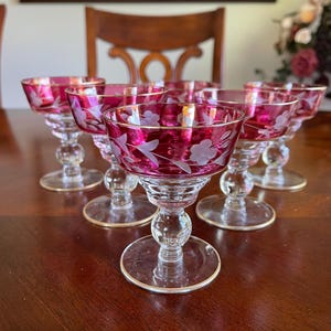 May include: Set of six vintage champagne glasses. Each glass features a ruby red bowl with etched floral designs and a gold rim. The glasses have a clear, round base and a decorative stem. The glasses are arranged on a dark wooden surface.