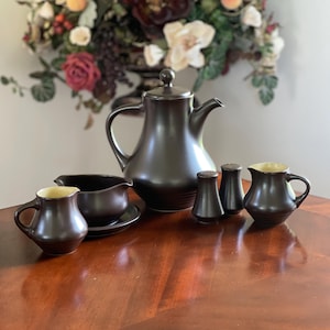 May include: A dark brown ceramic tea set featuring a large teapot with a lid, a creamer, a gravy boat on a small plate, and two matching salt and pepper shakers. Each piece has a smooth, matte dark brown exterior and a light cream-colored interior.