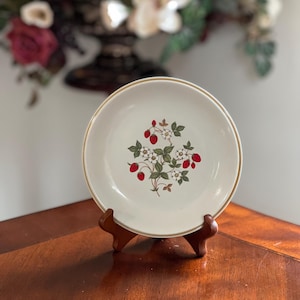 May include: A white ceramic plate with a gold rim and a strawberry design. The plate is supported by a wooden stand and is displayed on a wooden table. The strawberry design features red strawberries, white flowers, and green leaves.