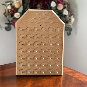 May include: A light brown, house-shaped wooden display board featuring a grid of circular indentations and small, clear plastic pegs protruding from the center of each circle, arranged in a symmetrical pattern across the flat surface.
