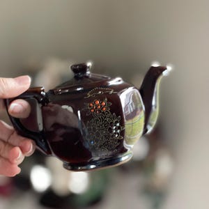 May include: A dark brown ceramic teapot with a lid, spout, and handle. The teapot is decorated with a floral design in gold and orange. The teapot is held in a person's hand.
