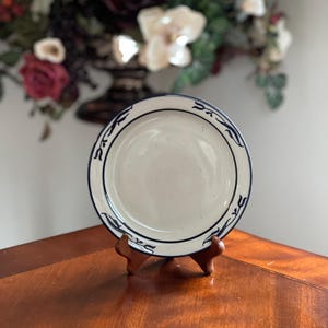 May include: A white ceramic plate with a blue decorative border and a dark blue rim. The plate is displayed on a wooden stand, showcasing its design. The plate's design features a repeating pattern of stylized leaves.