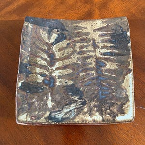 May include: Square ceramic plate with a unique, textured surface. The plate features a blend of brown, black, and blue hues, with a design reminiscent of foliage. The edges are slightly rounded, and the plate has a rustic, handmade appearance.